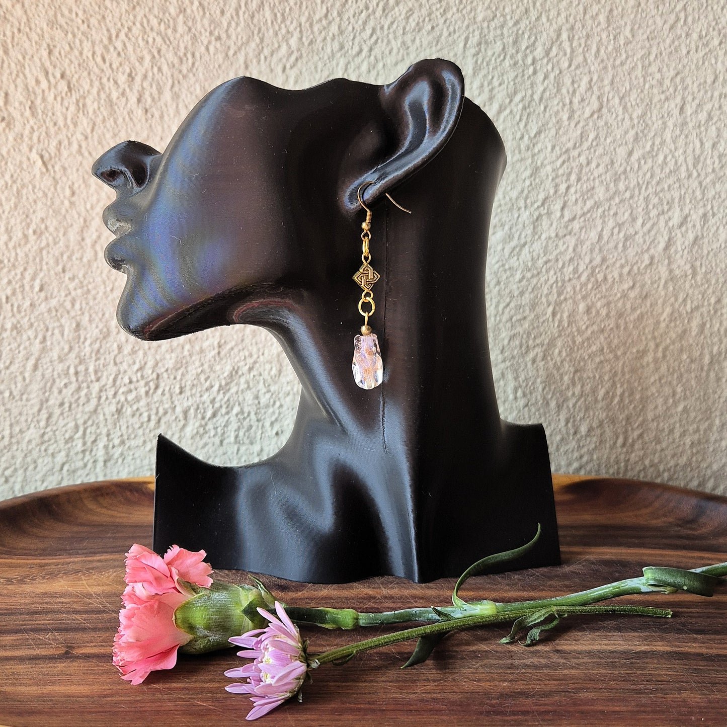 A single dangling earring displayed on a black mannequin head, with pink flowers on a wooden surface in front. The earring has a gold-toned geometric charm and a dangling faceted pale pink stone.