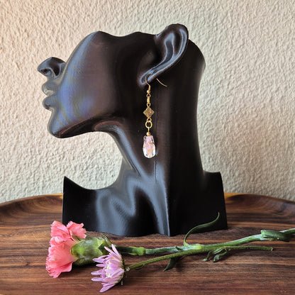 A single dangling earring displayed on a black mannequin head, with pink flowers on a wooden surface in front. The earring has a gold-toned geometric charm and a dangling faceted pale pink stone.