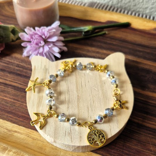 A gold-tone beaded bracelet with clear faceted beads and animal charms, including a tree of life pendant, displayed on a cat-shaped wooden stand.