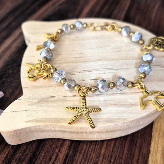 A gold-tone charm bracelet with faceted light blue beads, a starfish charm, and a small sea urchin charm, displayed on a light wood fish-shaped tray over a dark wood surface.