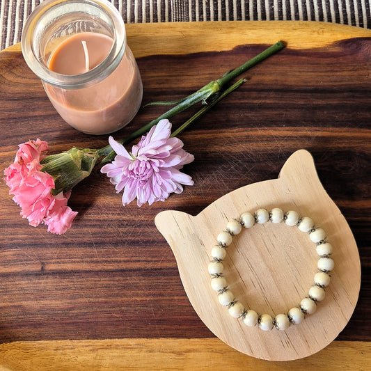 A beaded bracelet made of cream-colored round beads with small dark accents, displayed on a light wood cat-shaped stand on a dark wooden board, next to a coffee candle and two pink flowers.